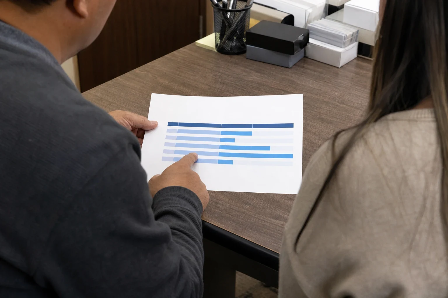 Over-the-shoulder view of a man pointing to a printed blue bar chart on a wooden desk while a woman looks on, with a pen cup and boxes in the background