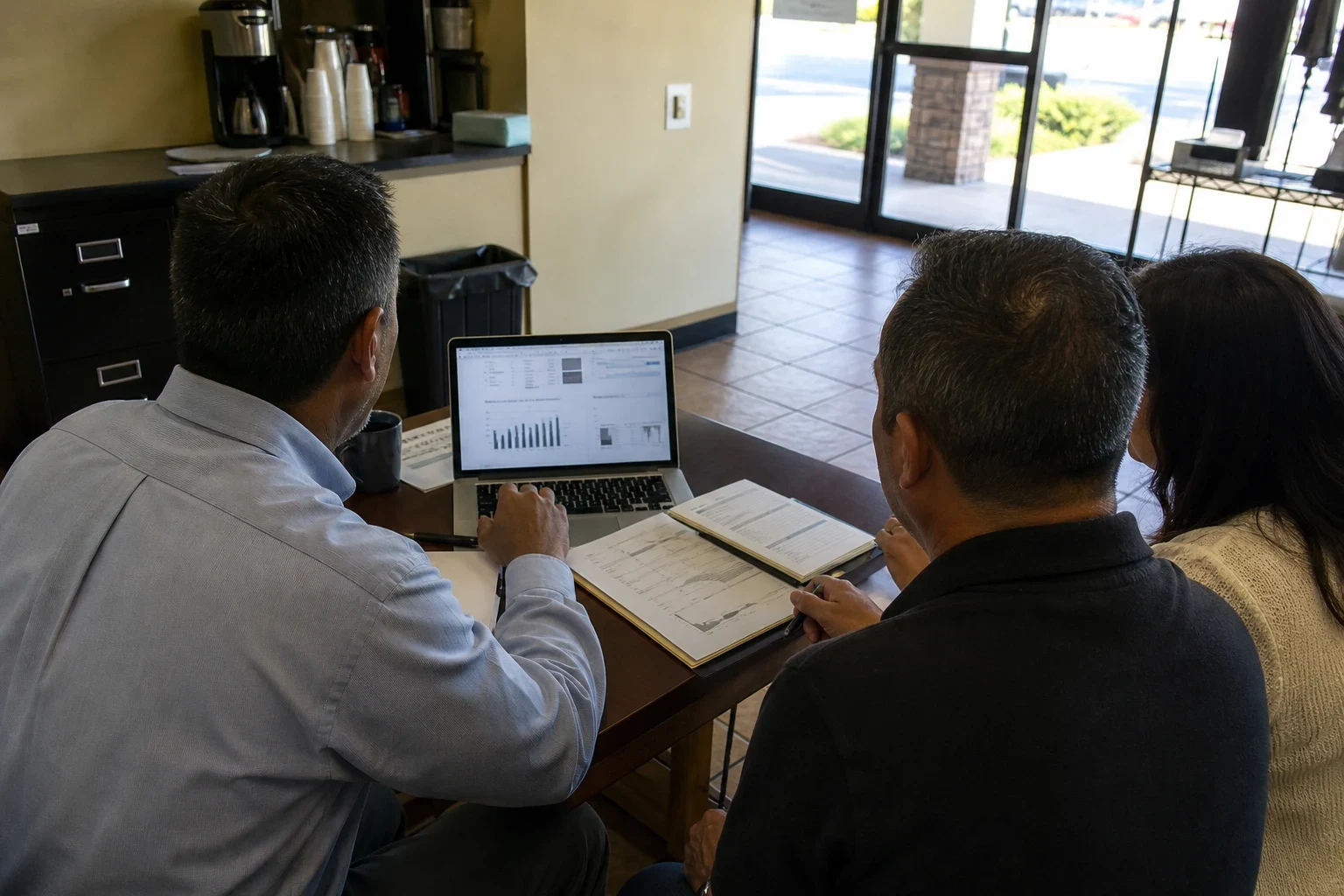 An agent in a light blue button-down shirt shows charts on a laptop to a man and woman at a small table in a storefront office with tile floor and glass doors